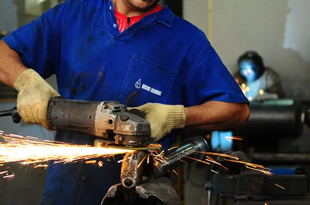 Operador de uniforme azul e luvas esmerilhando uma peça, com o jato de faíscas em primeiro plano
