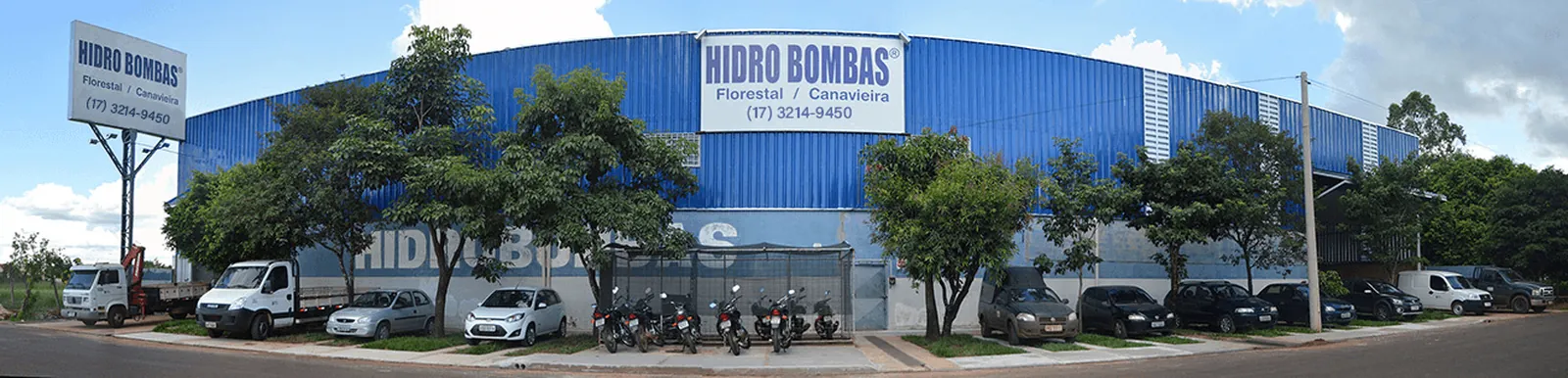 Fachada da Hidro Bombas: galpão de telha metálica azul com o letreiro HIDRO BOMBAS, Florestal / Canavieira, e carros estacionados ao longo da calçada
