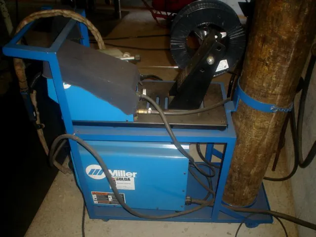 Máquina de solda Miller azul com o carretel de arame e o tocheiro, na área de solda