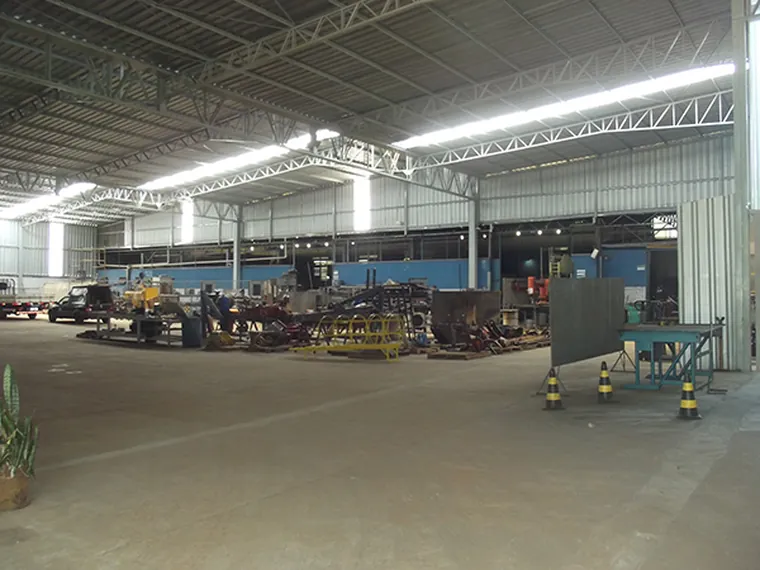 Salão interno da fábrica, com peças e equipamentos alinhados ao longo do vão livre sob o telhado metálico