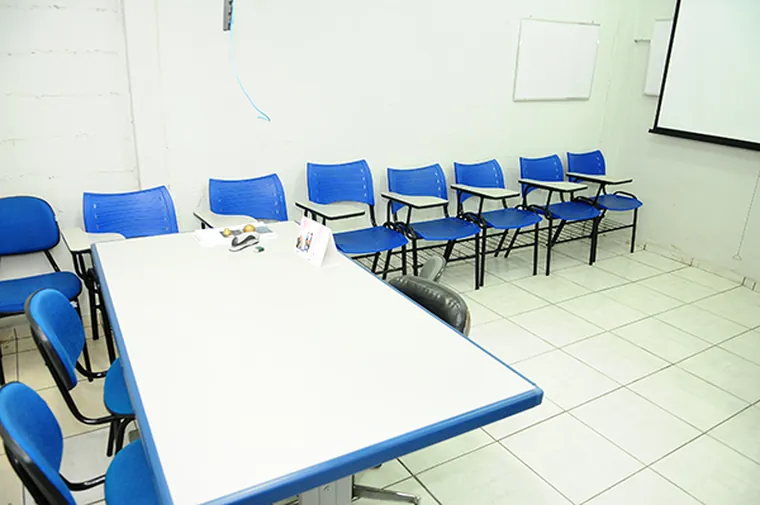 Sala de treinamento com mesa branca e cadeiras azuis voltadas para a tela de projeção
