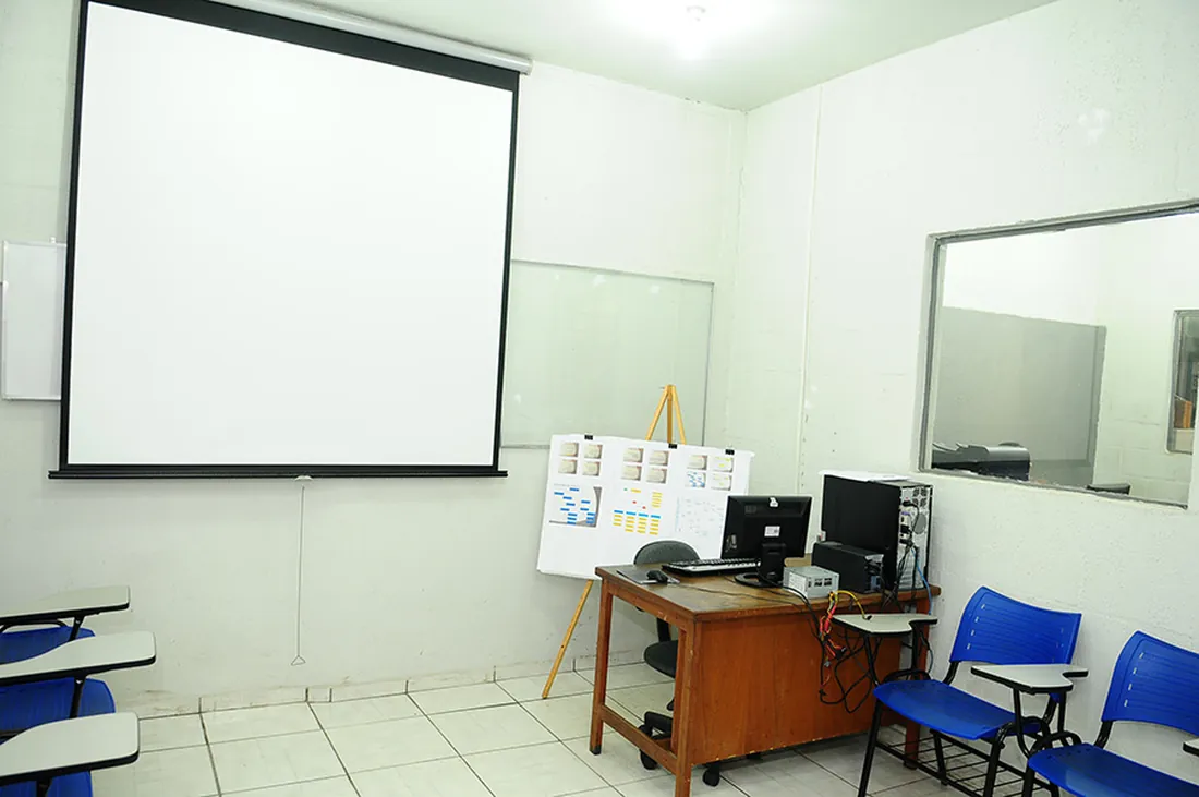 Sala de treinamento montada, com tela de projeção, computador e material didático sobre a mesa